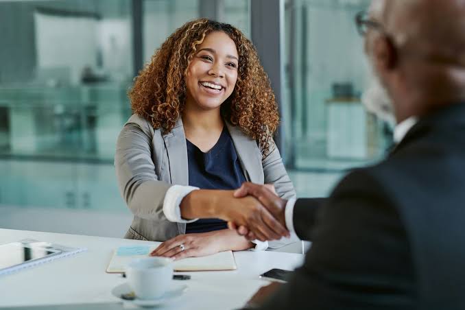 Professional recognition moment — a woman in a business meeting shaking hands with a senior executive across the table, representing earned visibility and career advancement